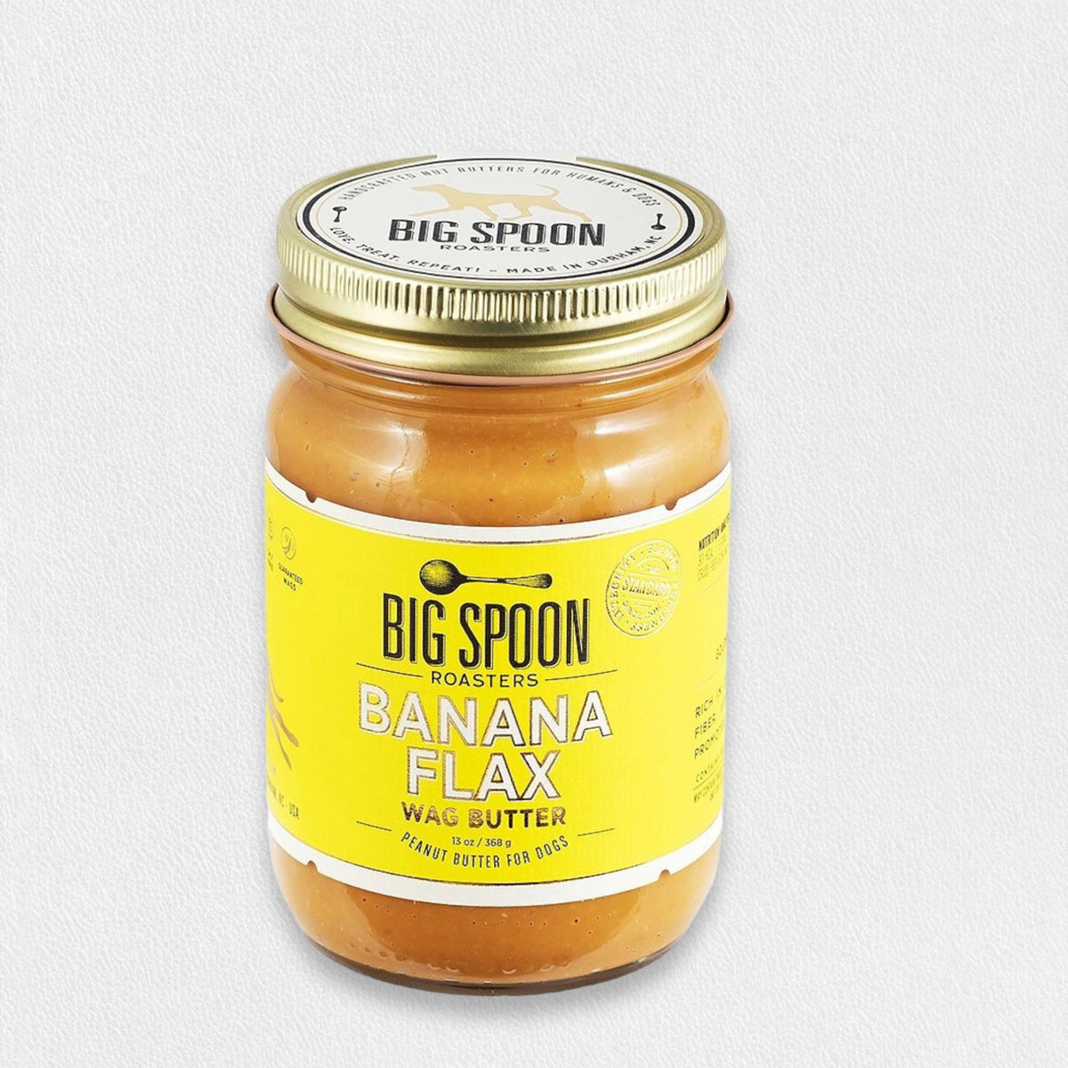 A jar of BIG SPOON ROASTERS - WAG BUTTER BANANA FLAX, a Canine Superfood rich in Omega-3 fatty acids, is shown against a plain white background. The jar features a yellow label with black text and a gold-colored lid, while the light brown butter inside resembles Peanut Butter for Dogs.