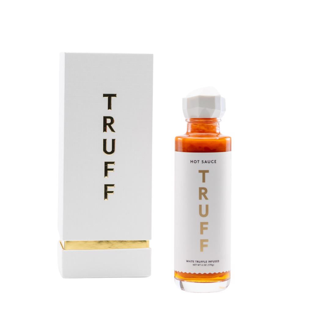A bottle of TRUFF - HOT SAUCE WHITE TRUFFLE is shown next to its white and gold rectangular packaging. Both the bottle and packaging prominently display the brand name "TRUFF HOT SAUCE" vertically in bold gold lettering, epitomizing a luxury hot sauce experience.
