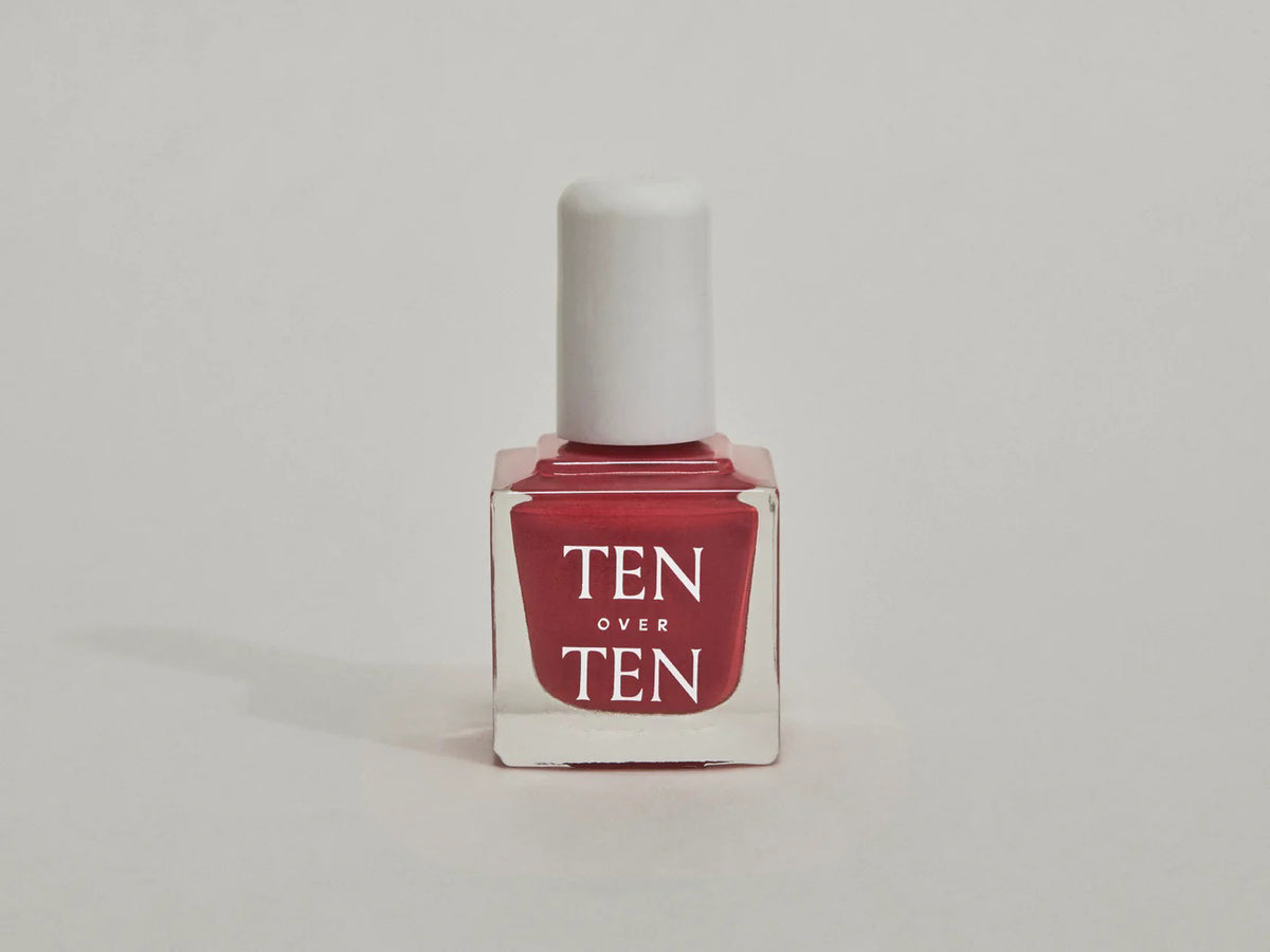 A glass bottle of TENOVERTEN - NAIL POLISH from the brand TEN OVER TEN, featuring a white cap and a dusty pink nail color. The front label displays "TEN OVER TEN" in white uppercase letters. This vegan, eight-free formula ensures both beauty and peace of mind. The background is plain and light-colored.