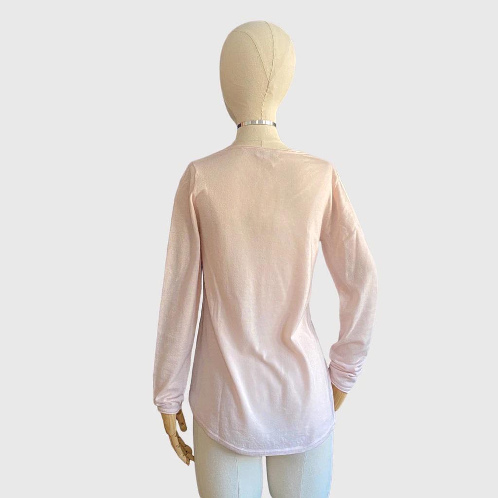A person wearing a shell pink Kinross Cashmere - Worsted Round Hem V-neck top and white pants is touching their hair with one hand. The background is plain white.