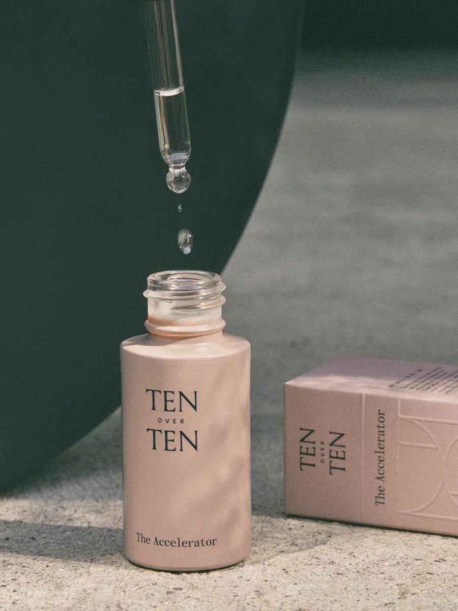 A glass dropper dispenses fast-acting serum into a pink bottle labeled "TENOVERTEN - THE ACCELERATOR QUICK DRY" by TEN OVER TEN, with a matching box beside it on a textured surface.