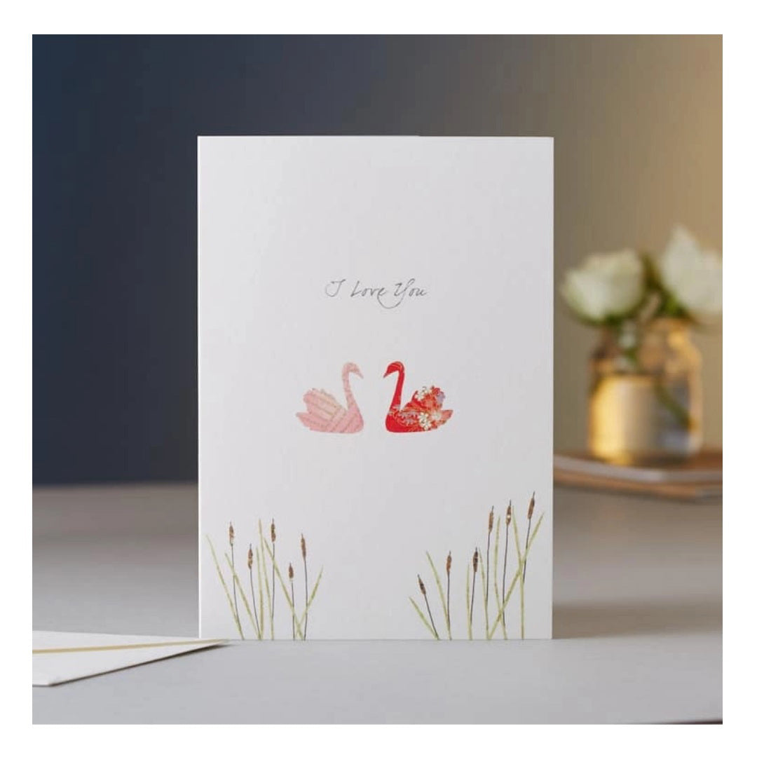 The ELOISE HALL - SWANS I LOVE YOU CARD by ELOISE HALL LTD is a luxury stationery greeting card featuring two swans, one pink and one red, with the words "I love you" written above them. The foreground includes green grass illustrations, while the softly lit background showcases a blurred vase with white flowers.