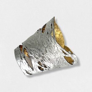 A person wearing the ILLUSIONISM BRACELET IN GOLD AND SILVER (REVERSIBLE) from SI SABATO ISABEL on their wrist. The handmade jewellery, crafted by an Argentinian artist, features irregular cut-out shapes and an intricate, hammered surface. The person is dressed in a faded blue denim shirt with white buttons.