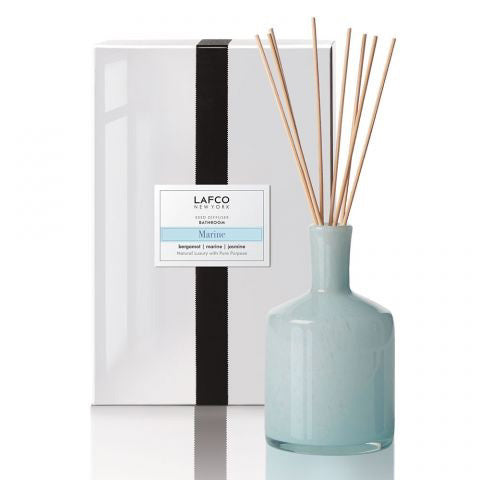 A light blue LAFCO - MARINE BATHROOM DIFFUSER bottle with multiple reed sticks is positioned in front of a white box adorned with a black vertical stripe. The box features the &quot;LAFCO New York&quot; label, and the diffuser is named &quot;Marine,&quot; combining notes of bergamot and sea jasmine for a refreshing coastal aroma.