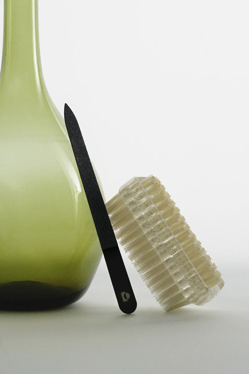 TENOVERTEN - THE SHAPER NAIL FILE by TEN OVER TEN and a translucent nail brush rest against a light green glass vase on a neutral backdrop.