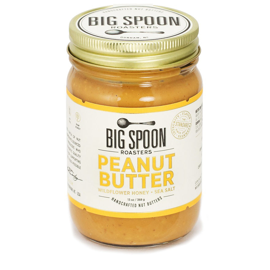 A jar of BIG SPOON ROASTERS - PEANUT WILDFLOWER HONEY NUT BUTTER is shown. The label mentions &quot;PEANUT WILDFLOWER HONEY NUT BUTTER,&quot; &quot;Wildflower Honey,&quot; &quot;Sea Salt,&quot; and &quot;Handcrafted Nut Butters.&quot; The lid is gold, and the packaging has a white background with yellow accents, highlighting the rich flavor profile of honey and sea salt.