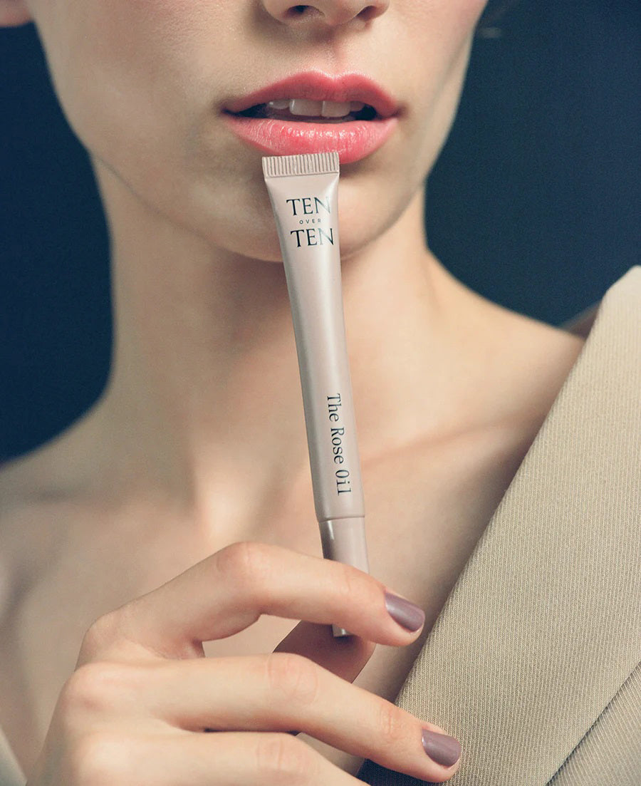 A tube and box of TEN OVER TEN - THE ROSE OIL CUTICLE TREATMENT stand upright on a dark, angular surface under soft lighting, emphasizing the brand’s hydrating formula for restoring nails and cuticles.
