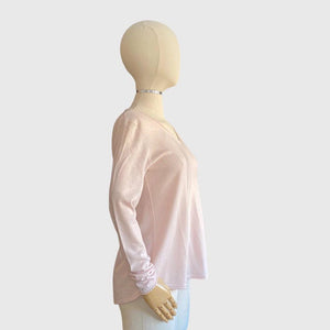 A person wearing a shell pink Kinross Cashmere - Worsted Round Hem V-neck top and white pants is touching their hair with one hand. The background is plain white.
