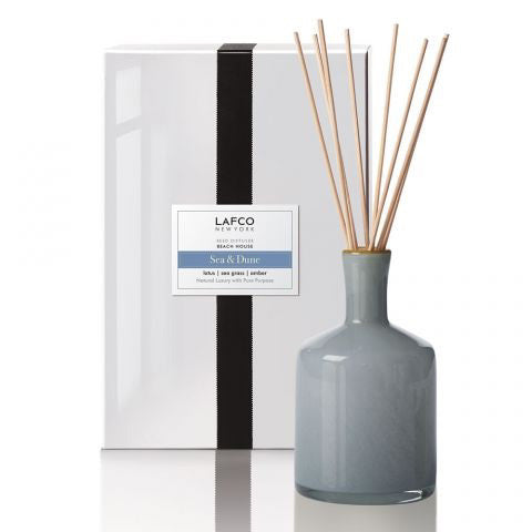A minimalist LAFCO SEA &amp; DUNE Beach House reed diffuser in a blue-gray bottle with natural reeds extending upward stands next to its packaging. The white box features a black vertical stripe and a label that reads "LAFCO New York - Beach House - Sea &amp; Dune." The elegant bottle exudes a clean aesthetic, reminiscent of serene coastal lilies.