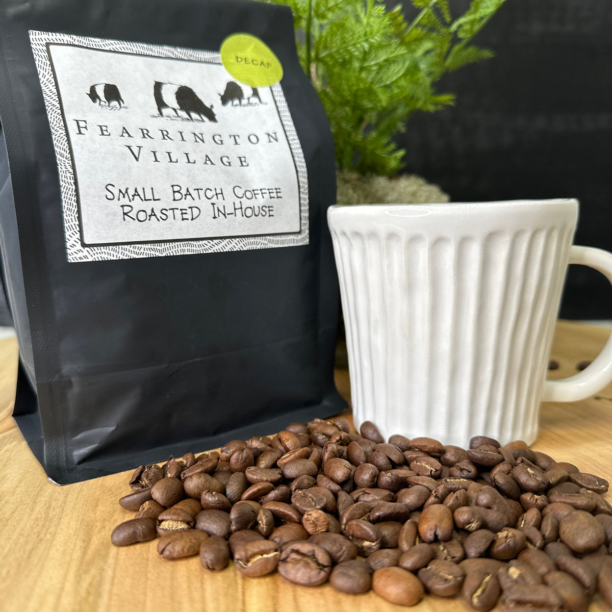 A black package of Belted Goat Fearrington Signature Collection Small Batch House Blended Decaf Coffee Beans. The label features three black animal silhouettes and a decorative border. A green sticker in the upper-right corner indicates the Fearrington House decaf blend, made from Brazilian and Colombian beans. The background is textured white.