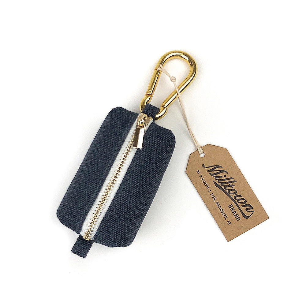 The DOG WASTE DISPENSER (MIDNIGHT) by MILLTOWN BRAND is a chic, small pouch made of navy blue pigment-washed canvas, featuring a white zipper and a gold-colored carabiner clip. A tan tag with &quot;Milltown Brand&quot; branding hangs from the carabiner.