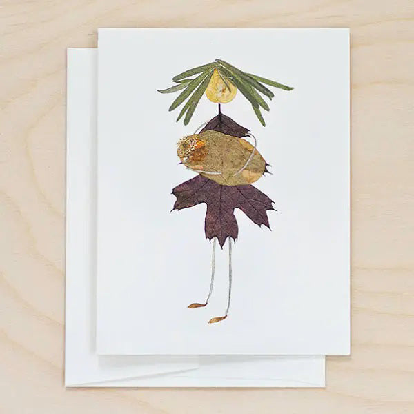Celebrate Mother's Day with the PETAL PEOPLE PRESS - MOM CARD by PETAL PEOPLE PRESS, featuring a delightful figure crafted from pressed botanicals. The head is a small, round leaf, the body showcases an array of colorful leaves, and the arms and legs are depicted with slender plant stems. This charming card rests on a light wooden surface.