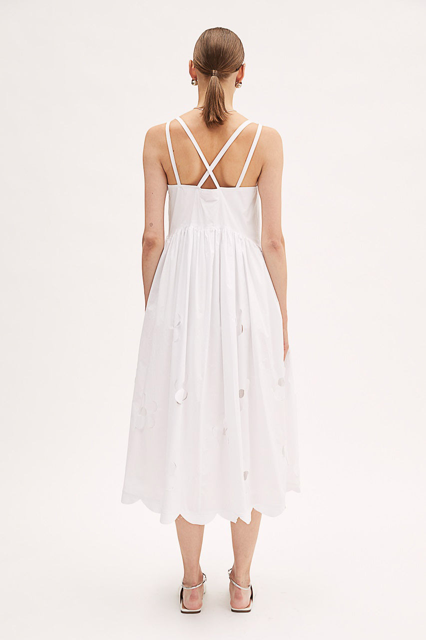 A person wearing the MEIMEIJ Cotton Midi Dress, featuring sleeveless design with delicate cut-out floral patterns, stands against a plain white background. The light dress from MEIMEIJ includes criss-cross straps, a gathered waist, and a scalloped hemline. They are also wearing white shoes with pointed toes.