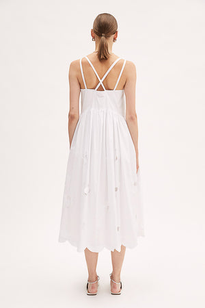 A person wearing the MEIMEIJ Cotton Midi Dress, featuring sleeveless design with delicate cut-out floral patterns, stands against a plain white background. The light dress from MEIMEIJ includes criss-cross straps, a gathered waist, and a scalloped hemline. They are also wearing white shoes with pointed toes.