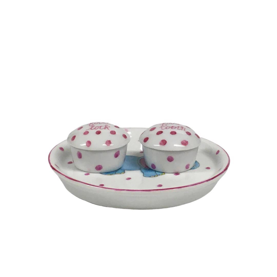 Introducing the TOOTH AND LOCK ELEPHANT (PINK) by ANN MARIE MURRAY, L.L.C., a bespoke porcelain set that includes a beautifully crafted plate and two small lidded containers. The plate showcases a hand-painted blue elephant with pink ears playing with a colorful ball, all surrounded by charming pink polka dots. The dotted lidded containers are thoughtfully labeled "first lock" and "first tooth.
