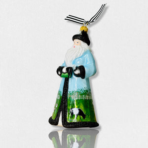 The Fearrington Signature Collection - Handmade Santa Ornament by Joy to the World Collectibles features a bearded figure in a blue robe adorned with farm scenes, including sheep, chickens, and a white fence. This hand-painted keepsake from Fearrington Village is topped with a gold cap and includes a black-and-white striped ribbon for hanging.
