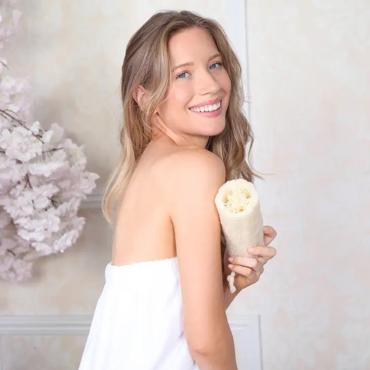 Image of the DAILY CONCEPTS BODY LOOFAH product in its packaging. The white packaging displays an image of the loofah, text describing it as a gentle exfoliation tool, and logos signifying its organic and vegan qualities. The biodegradable, rectangular loofah has a coarse texture.