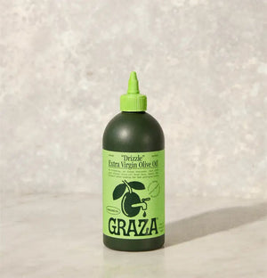 A green squeeze bottle labeled "DRIZZLE OLIVE OIL" by GRAZA sits on a gray surface against a light gray textured background. The bottle features green and black graphics, including an olive branch, making it perfect for adding flavor as a finishing oil.