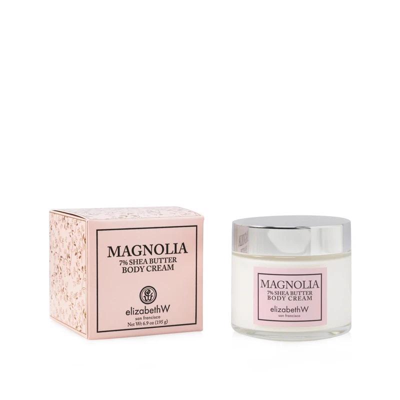 A jar of MAGNOLIA BODY CREAM from E WIGHTMAN &amp; CO is displayed next to its product box. The box is light pink with decorative floral accents, while the jar, holding an intensely hydrating and indulgent cream, has a silver lid and a matching pink label.