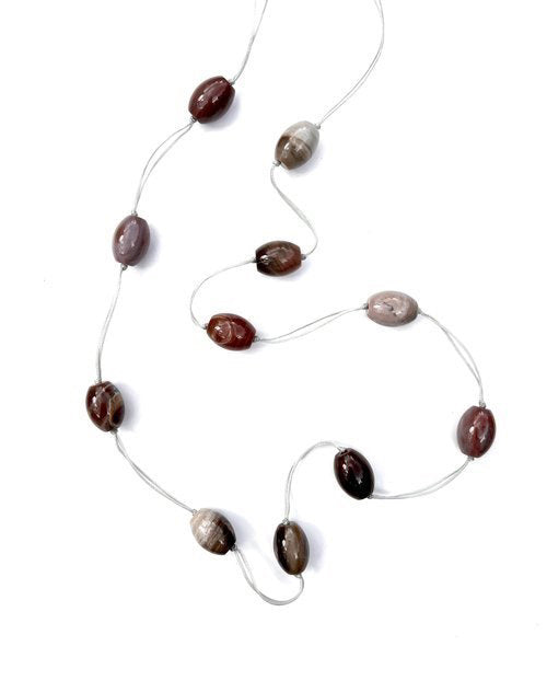 The ANN LIGHTFOOT - INTERMITTENT STRAND NECKLACE OF PETRIFIED WOOD by ANN LIGHTFOOT features an elegant 35” strand of oval brown, reddish, and gray beads strung on a thin, transparent thread. Each bead slightly varies in color and pattern, enhancing its natural aesthetic. Barrel-shaped beads are interspersed throughout to add texture.