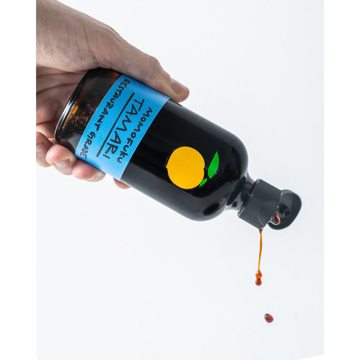 A brown glass bottle with a black cap contains MOMOFUKU - TAMARI, a restaurant-grade, gluten-free soy sauce made from organic ingredients. The label is blue with a minimalist orange fruit logo. The bottle is reflected on the white surface it sits on.