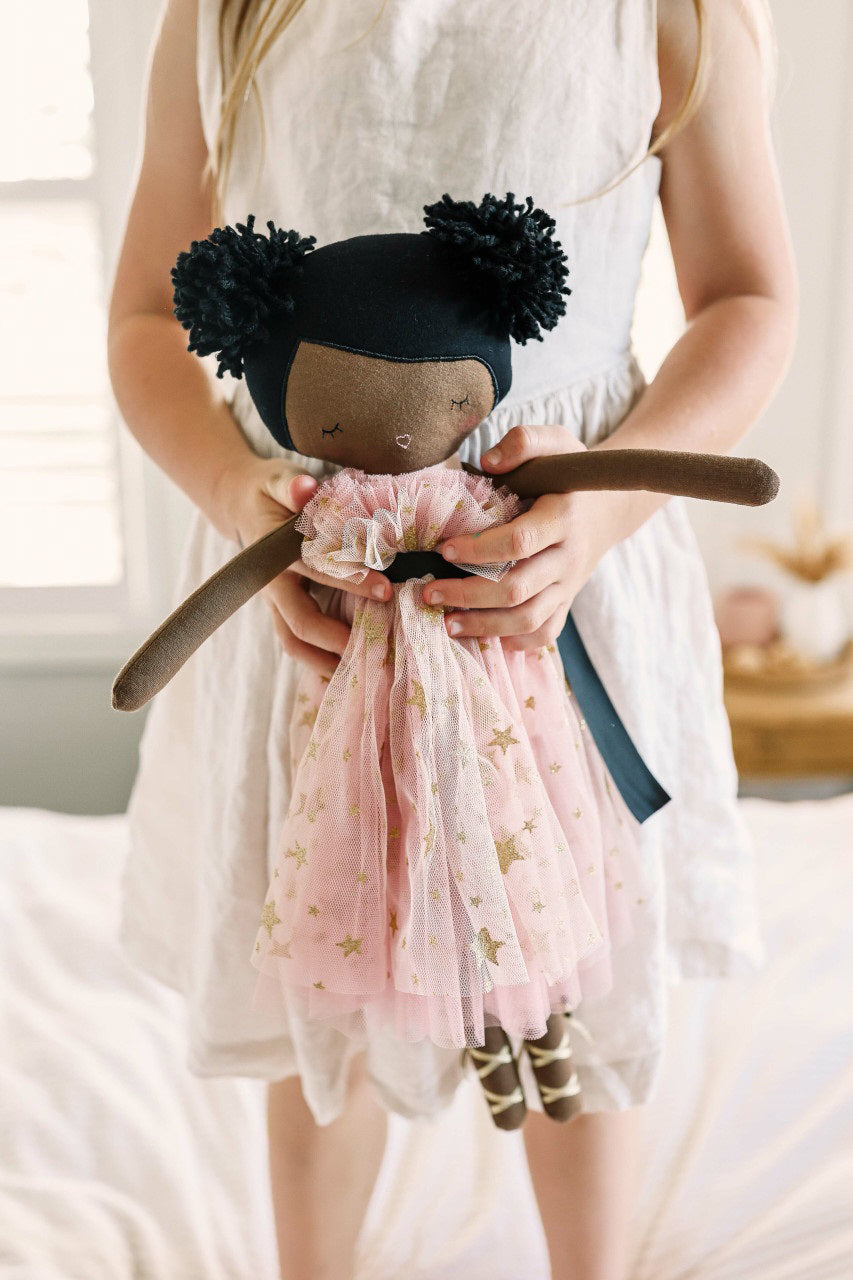 The ALIMROSE MAE Doll Blush Gold Star by ALIMROSE features a handmade design with brown skin and adorable pom-pom hair styled in two puffs. She is dressed in a blush pink tulle dress adorned with gold stars, cinched at the waist with a black ribbon. She completes her enchanting look with ballet-style shoes and an embroidered face accentuated by charming details.