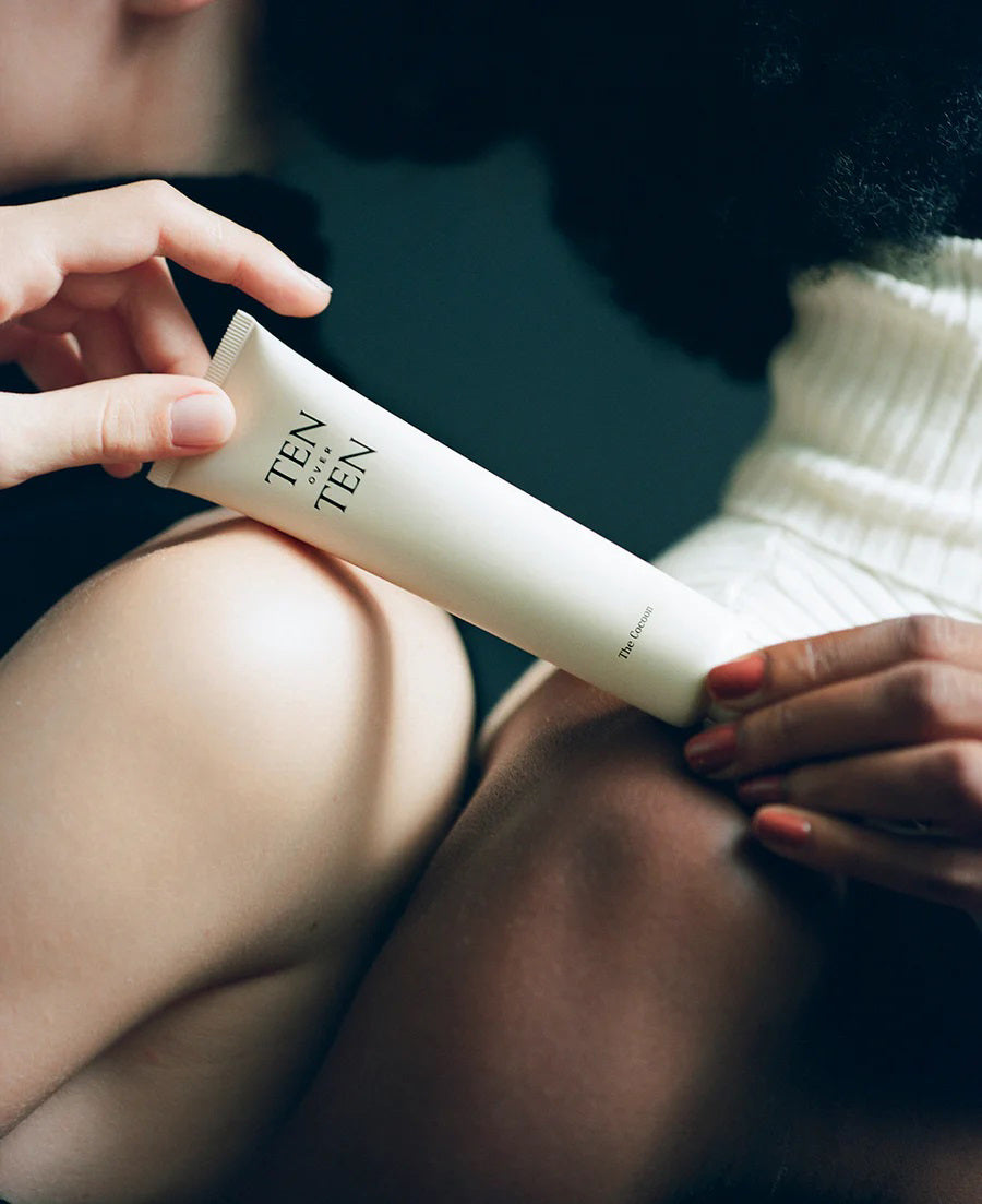 Two people, one with bare shoulders and another in white, hold a TENOVERTEN - THE COCOON HAND &amp; FOOT CREAM tube by TEN OVER TEN—suggesting a plant-based cream for long-lasting moisture on dry hands and feet.