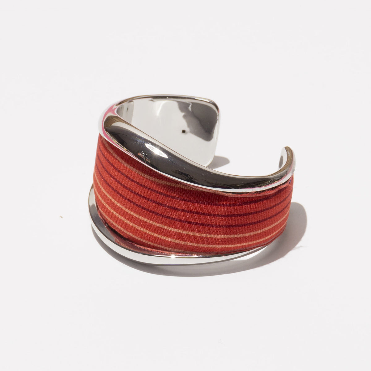 Discover the VINTAGE SCARF BANGLE (RED) by MILANO MM33—a stylish cuff bracelet with a wide band framed in glossy silver. Adorned with gradient red and dark orange striped fabric made from vintage silk, it offers a vibrant touch of color. The interior is smooth and reflective, ensuring comfort and elegance. Proudly MADE IN ITALY against a plain white background.