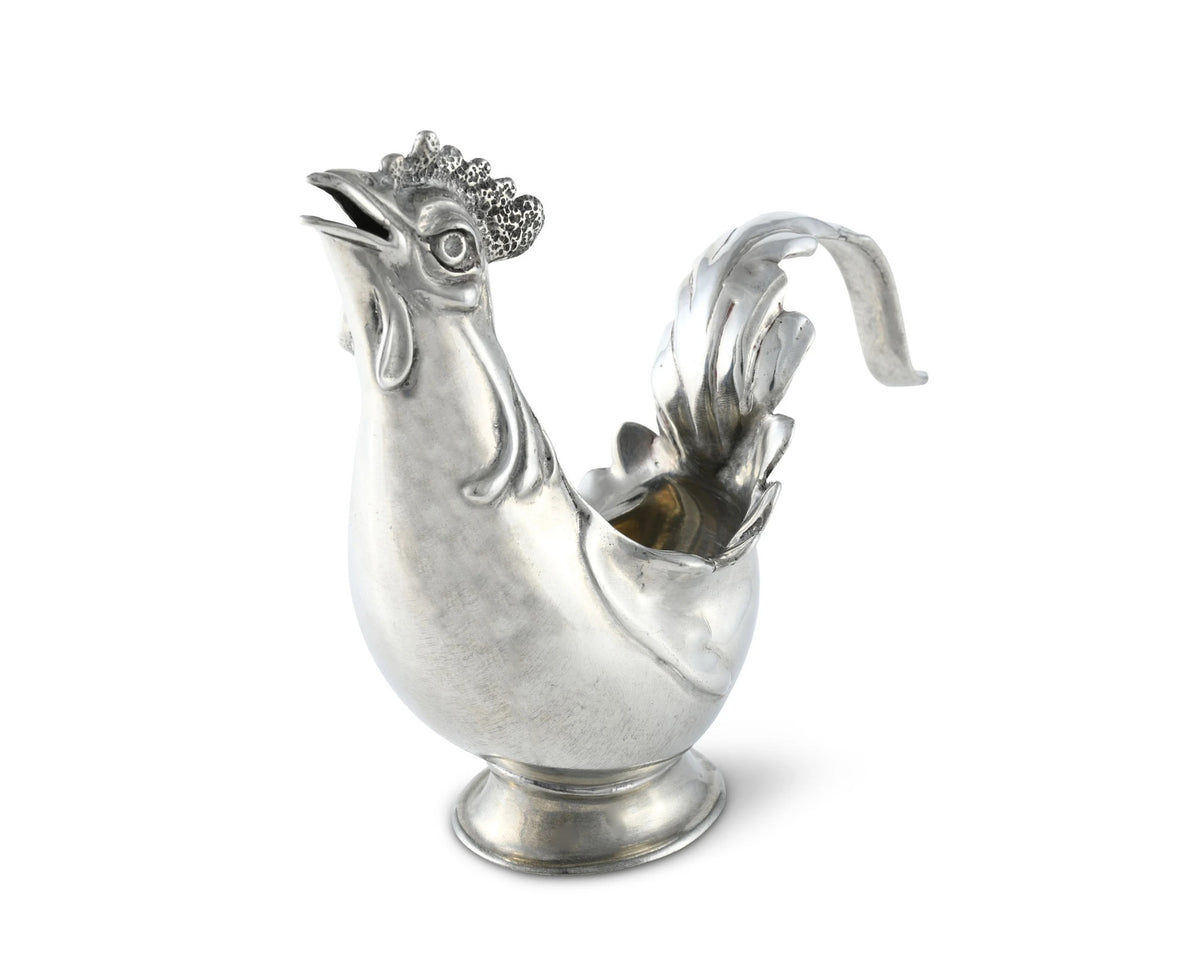 The VAGABOND HOUSE - PEWTER ROOSTER CREAMER features a rooster shape with an open beak spout, an ornate tail-turned handle, and detailed comb and feathers. Its handcrafted pewter craftsmanship ensures a shine on the round base, reflecting intricate details similar to premium tableware.
