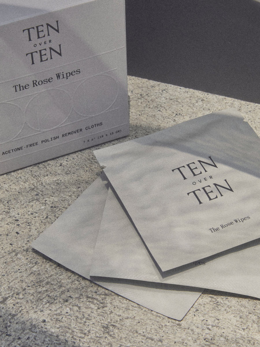 Three packets of TENOVERTEN - THE ROSE WIPES POLISH REMOVER by TEN OVER TEN are placed beside a matching box on a textured surface. These acetone-free wipes are ideal for removing nail polish while traveling.