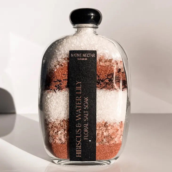 A glass bottle filled with layered bath salts in shades of pink, white, and brown. The black label reads "NATIVE NECTAR" at the top and "HIBISCUS &amp; WATERLILY FLORAL SALT SOAK" vertically down the side, showcasing its mineral-rich sea salt properties. The bottle is set against a plain background.