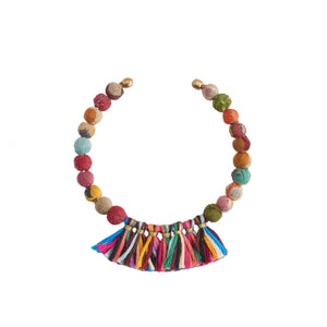 A colorful beaded bracelet lies on a white speckled plate. The KANTHA FRINGE CUFF from WORLD FINDS features small round beads in various bright colors and a vibrant tassel made of multicolored threads, forming a semicircle pattern. A green leaf partially overlaps the plate in the background, enhancing its boho look.