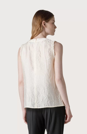 A person with long, straight hair wears a sleeveless, textured SLEEVELESS TOP by SEVENTY and black pants. They are posing against a plain, light grey background. The top features raw-cut armholes and a crinkled appearance, adding depth and visual interest to the outfit.