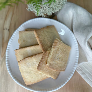 A stack of buttery and crisp vanilla shortbread cookies is displayed on a black serving board in front of a clear package labeled "Fearrington House Signature Collection - Housemade Vanilla Shortbread Cookies" with a black and white striped ribbon tie at the top. These delectable treats could easily be found at The Fearrington House Restaurant.