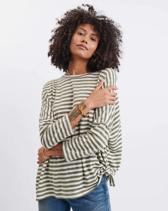 A person with curly hair wears a long-sleeved, green and white striped top with a side tie and blue jeans, standing against a plain white background.