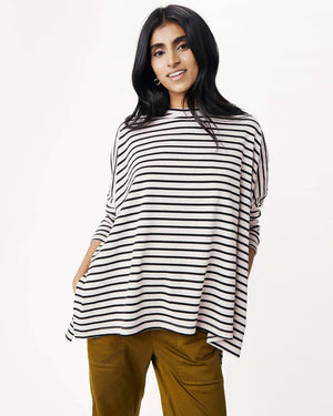 A woman with long, straight hair stands against a white wall, wearing the MERSEA Catalina Slub Tee in blue paired with white pants. The midweight textured cotton tee is from MERSEA. She looks at the camera with a neutral expression.