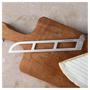 A handmade PRO SOFT CHEESE KNIFE with a cut-out design and the brand name "FORMATICUM" etched on its stainless steel blade is placed on a wooden cutting board. A partially sliced piece of cheese rests next to this professional cheese knife, crafted in Italy.