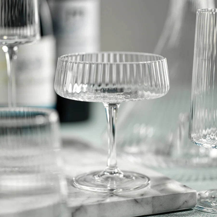 The FLUTED TEXTURED MARTINI GLASS by ZODAX stands empty against a white background, showcasing its wide, shallow bowl perched on a thin stem. Vertical ridges adorn the clear surface of the glass, adding texture to its elegant design. A flat, round base ensures stability.