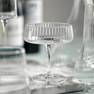The FLUTED TEXTURED MARTINI GLASS by ZODAX stands empty against a white background, showcasing its wide, shallow bowl perched on a thin stem. Vertical ridges adorn the clear surface of the glass, adding texture to its elegant design. A flat, round base ensures stability.