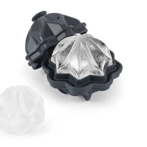 A black, hexagonal W&P - Ripple Cocktail Ice Mold with its top half removed, revealing a clear ice cube inside shaped like a decorative sphere with petal-like engravings. Another identical ice cube is shown next to the mold, perfect for chilling drinks with its intricate sculptural wave pattern from W&P Design.