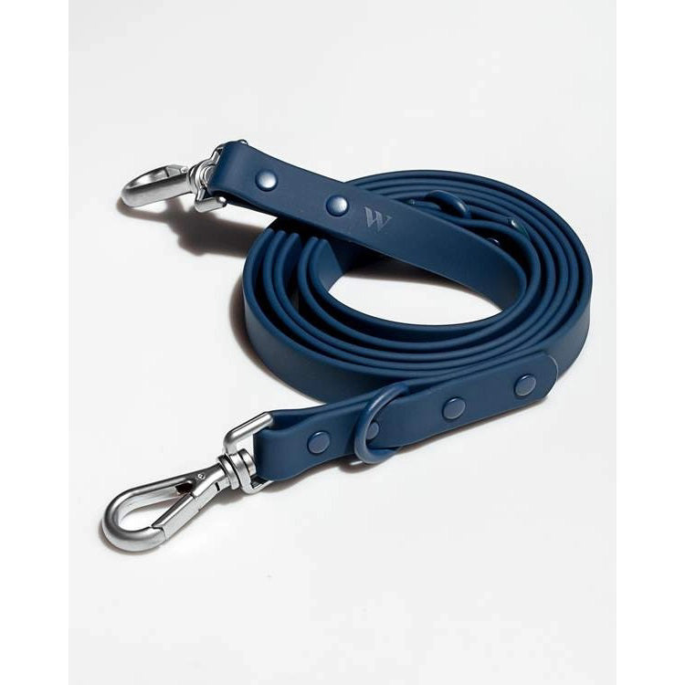 A coiled navy DOG LEASH STANDARD from WILD ONE with metal clips at both ends lays on a white surface. The leash, made from dirt and odor-resistant flex-poly straps, features a sleek design with rivet details and an embossed &quot;W&quot; near the top clip.