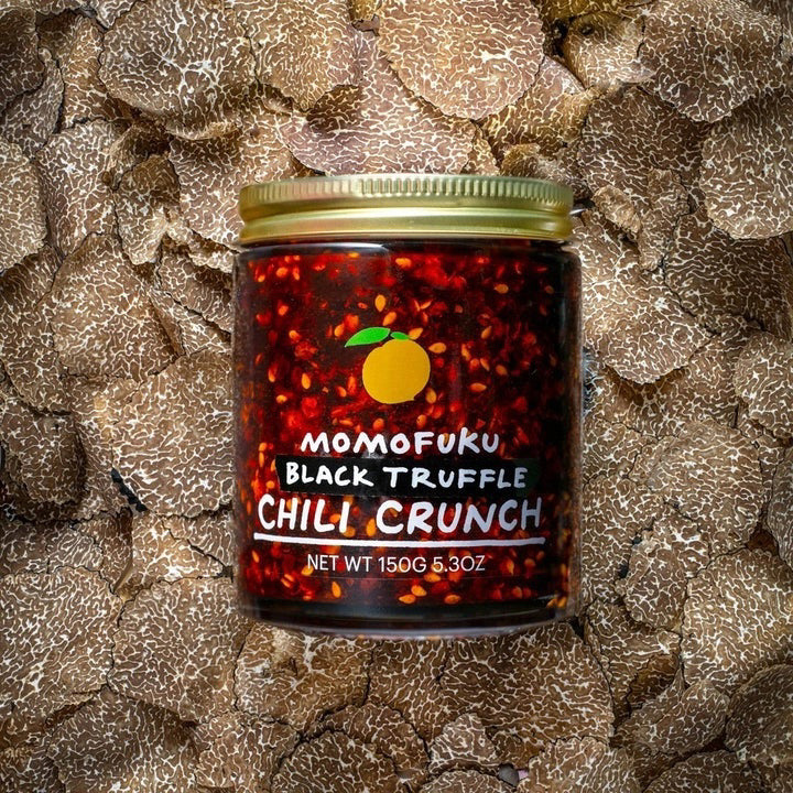 A jar of MOMOFUKU - BLACK TRUFFLE CHILI CRUNCH is shown against a plain background. The jar contains a mixture of chili flakes and oil, and features a label with an orange logo and text indicating "MOMOFUKU - BLACK TRUFFLE CHILI CRUNCH" alongside the net weight of 150g (5.3 oz).