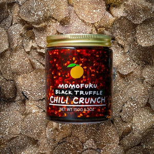 A jar of MOMOFUKU - BLACK TRUFFLE CHILI CRUNCH is shown against a plain background. The jar contains a mixture of chili flakes and oil, and features a label with an orange logo and text indicating "MOMOFUKU - BLACK TRUFFLE CHILI CRUNCH" alongside the net weight of 150g (5.3 oz).
