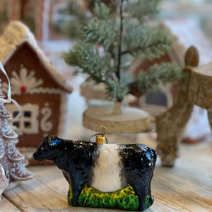 A luminous, hand-painted FEARRINGTON SIGNATURE COLLECTION - HANDMADE BELTIE ORNAMENT from JOY TO THE WORLD COLLECTIBLES dangles by a black and white striped ribbon. Shaped like a cow with a glossy surface that catches the light, it features a black head and rear with a white midsection. This delightful piece exudes the spirit of Fearrington Village, perched on a green base resembling grass.