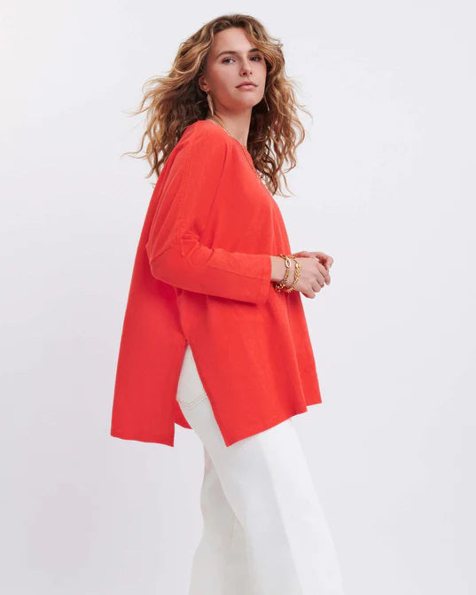 A woman with long, straight hair stands against a white wall, wearing the MERSEA Catalina Slub Tee in blue paired with white pants. The midweight textured cotton tee is from MERSEA. She looks at the camera with a neutral expression.