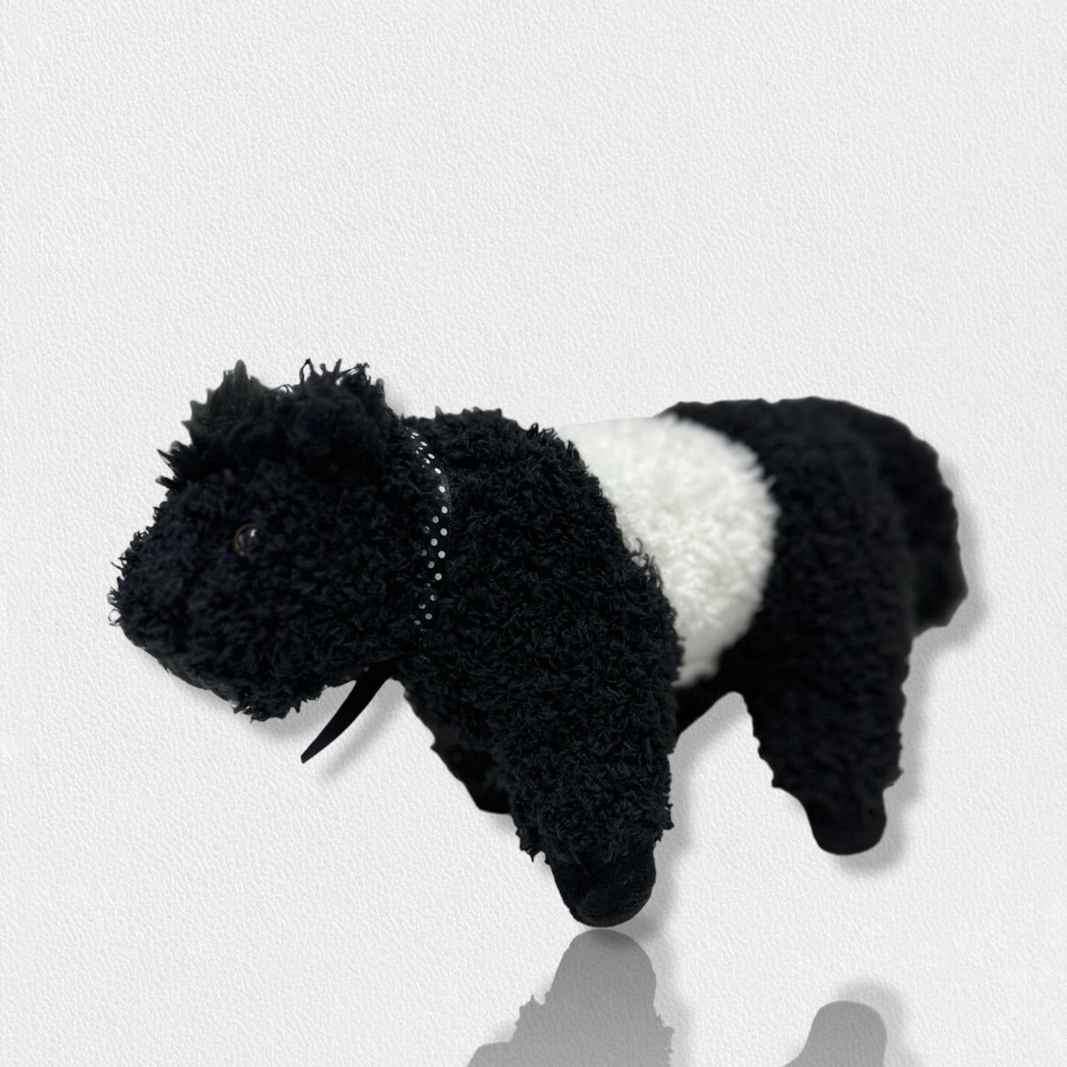 A soft plush toy from the Beverly Hills Teddy Bear Co&#39;s Fearrington Signature Collection, resembling a black and white Belted Galloway cow, stands against a plain white background. This furry mascot features black fuzzy fur on its head, legs, and tail, with a white fuzzy band around its midsection, characteristic of the Galloway breed.