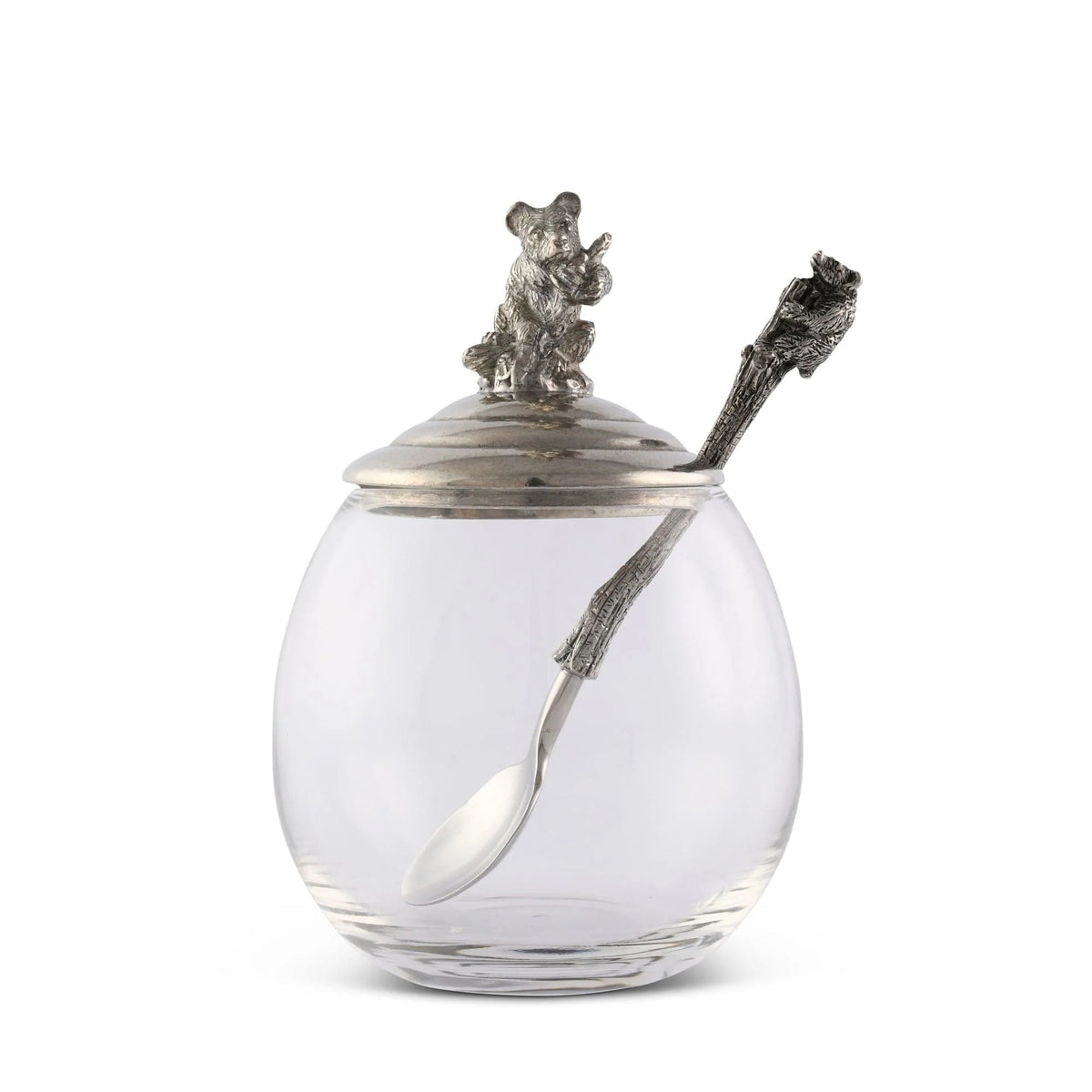 The VAGABOND HOUSE - LITTLE BEAR HONEY POT is a clear glass jar with a luxury pewter lid featuring a bear on top and includes a tree branch-styled silver spoon. This elegant yet rustic design by VAGABOND HOUSE resembles a little bear honey pot.