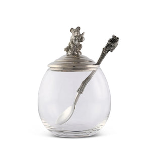 The VAGABOND HOUSE - LITTLE BEAR HONEY POT is a clear glass jar with a luxury pewter lid featuring a bear on top and includes a tree branch-styled silver spoon. This elegant yet rustic design by VAGABOND HOUSE resembles a little bear honey pot.