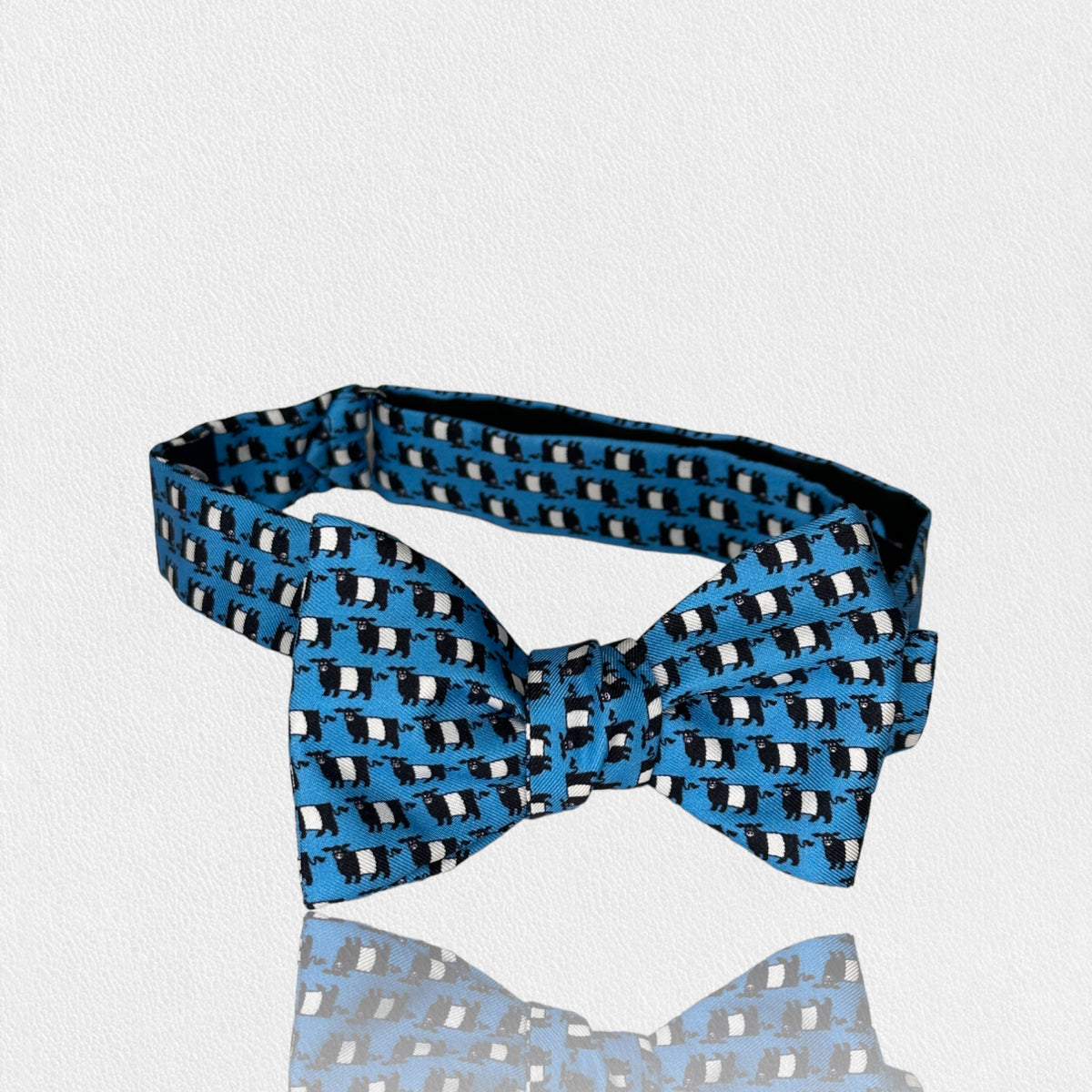 The FEARRINGTON SIGNATURE COLLECTION - BESPOKE BELTIE BOW TIE IN BLUE by COLLARED GREENS is a stylish accessory featuring a repeating pattern of small upright black and white penguins. Handmade from silk, this playful yet elegant bow tie is perfect for both formal and whimsical occasions, making it an excellent addition to any men&#39;s accessory collection.