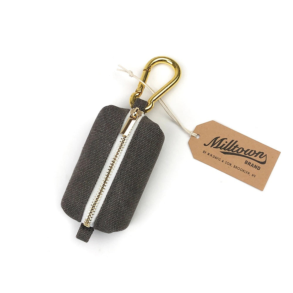 A compact, graphite-colored fabric pouch crafted from waxed canvas featuring a zippered closure and a gold-colored metal carabiner attachment. The pouch includes a tan tag displaying &quot;Milltown Brand by Bender &amp; Loin, Brooklyn, NY&quot; hanging from the clip. Perfect as an elegant dog waste bag dispenser.